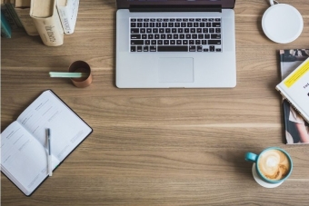 A wooden desk top with a planner, books, cup of coffee, and a laptop